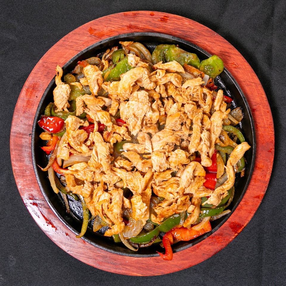 Sizzling skillet of seasoned sliced chicken with green and red peppers, onions, and mushrooms on a red wooden serving board.