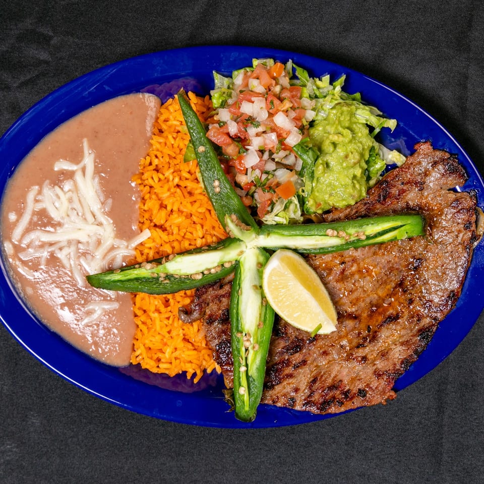 Mexican-style platter with grilled steak, orange rice, refried beans, salad, guacamole, pico de gallo, and lime on a blue plate.