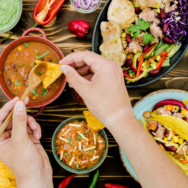 Overhead view of hands dipping tortilla chips into chili, surrounded by Mexican dishes.